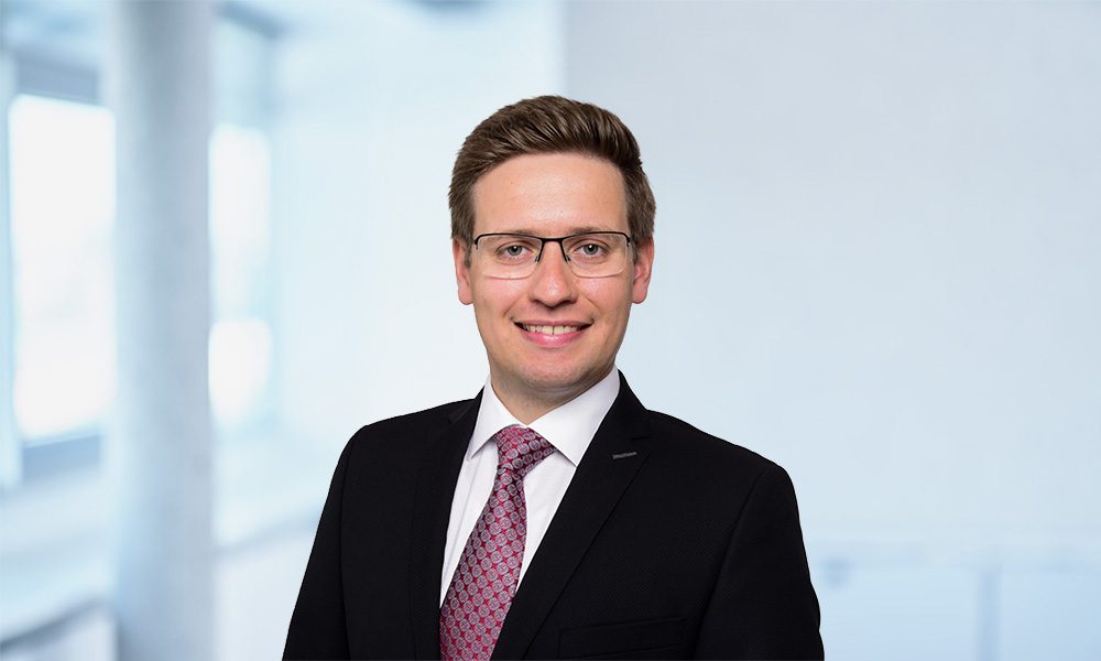 Maximilian Trappmann_web Smiling businessman in a black suit and pink tie, wearing glasses, in a bright office setting.