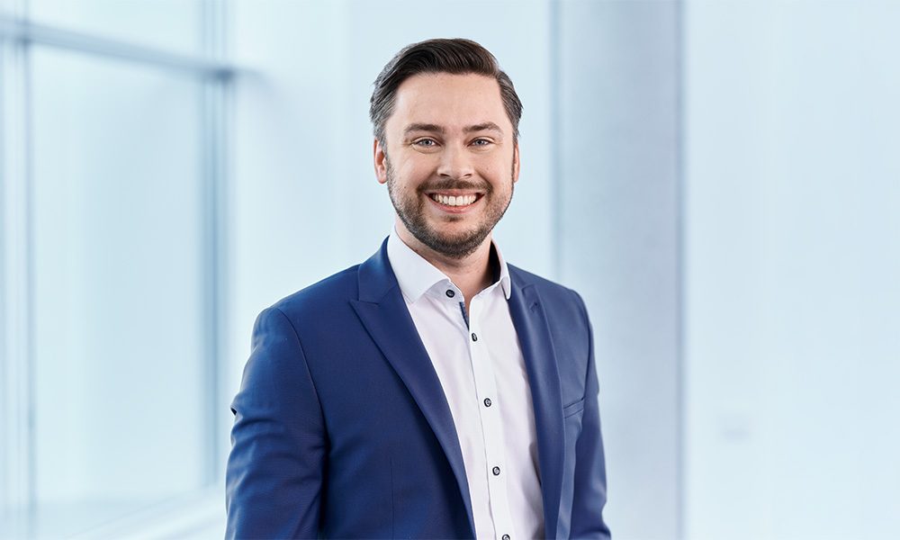 Andreas Spindler_web Smiling man in a navy suit and white shirt standing in a bright, modern office.