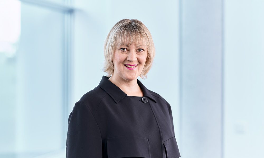 Franziska Kristin Grafe_web Professional portrait of a smiling woman with blond hair in a black blazer, set against a bright office background.