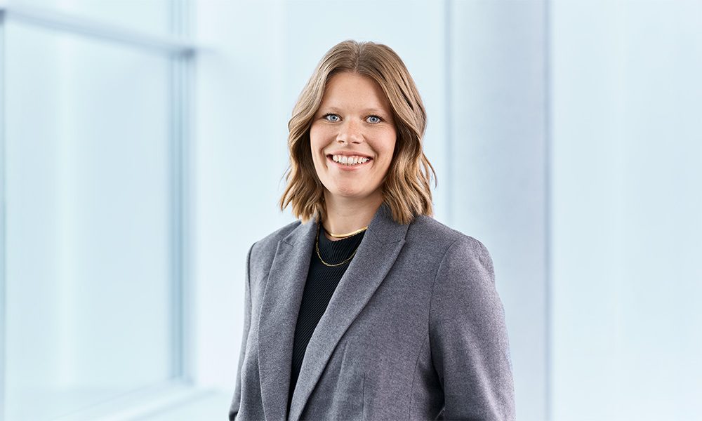 Sofia Mühlberger_web Professional woman in a gray blazer, smiling in a bright office for a corporate headshot.