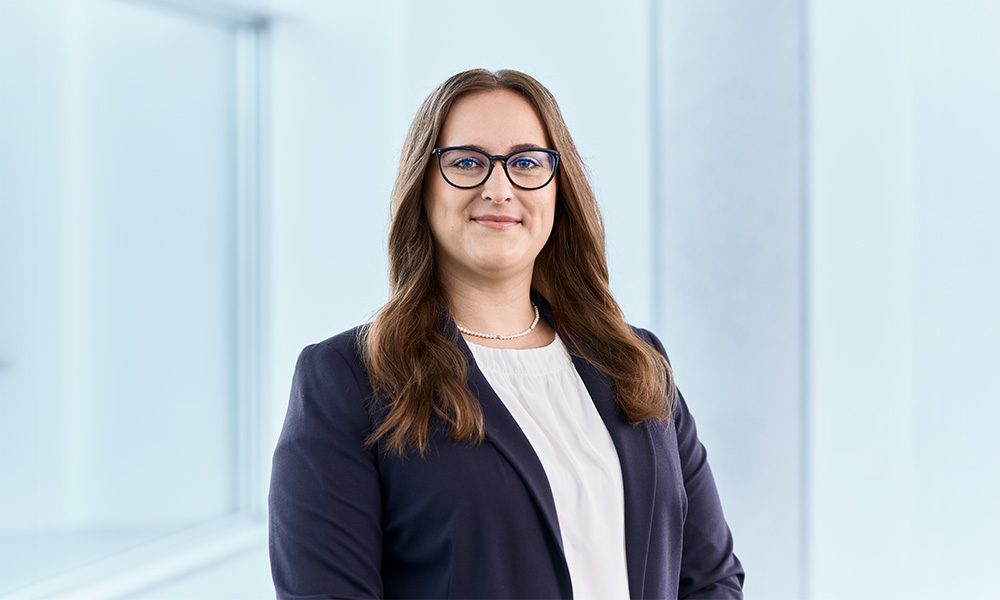 Melanie Volz_web Professional woman with glasses in a navy blazer and white blouse standing in a bright office.