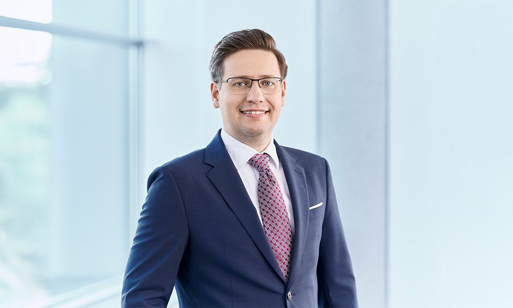 Maximilian Trappmann_01_web Smiling businessman in a black suit and pink tie, wearing glasses, in a bright office setting.