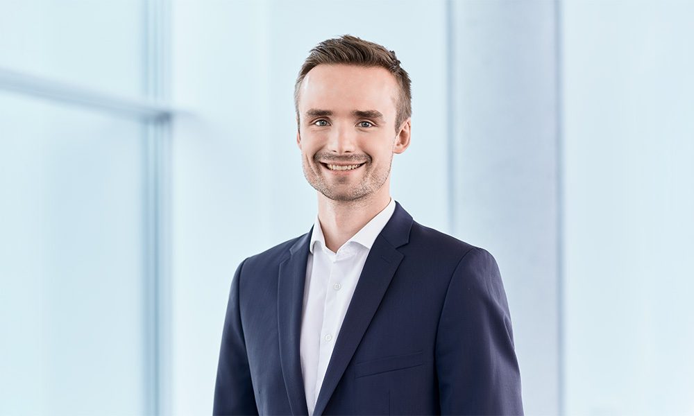 Lucas Plank_web Professional headshot of a smiling man in a navy suit and white shirt against a bright office backdrop.